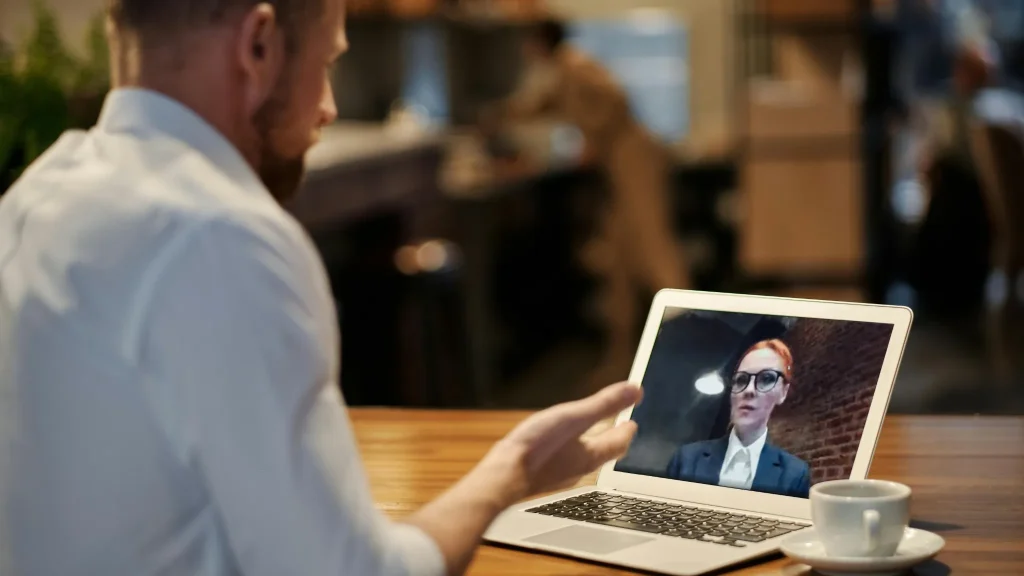 Mężczyzna przy stoliku w kawiarni rozmawia przez wideo z kobietą widoczną na ekranie laptopa